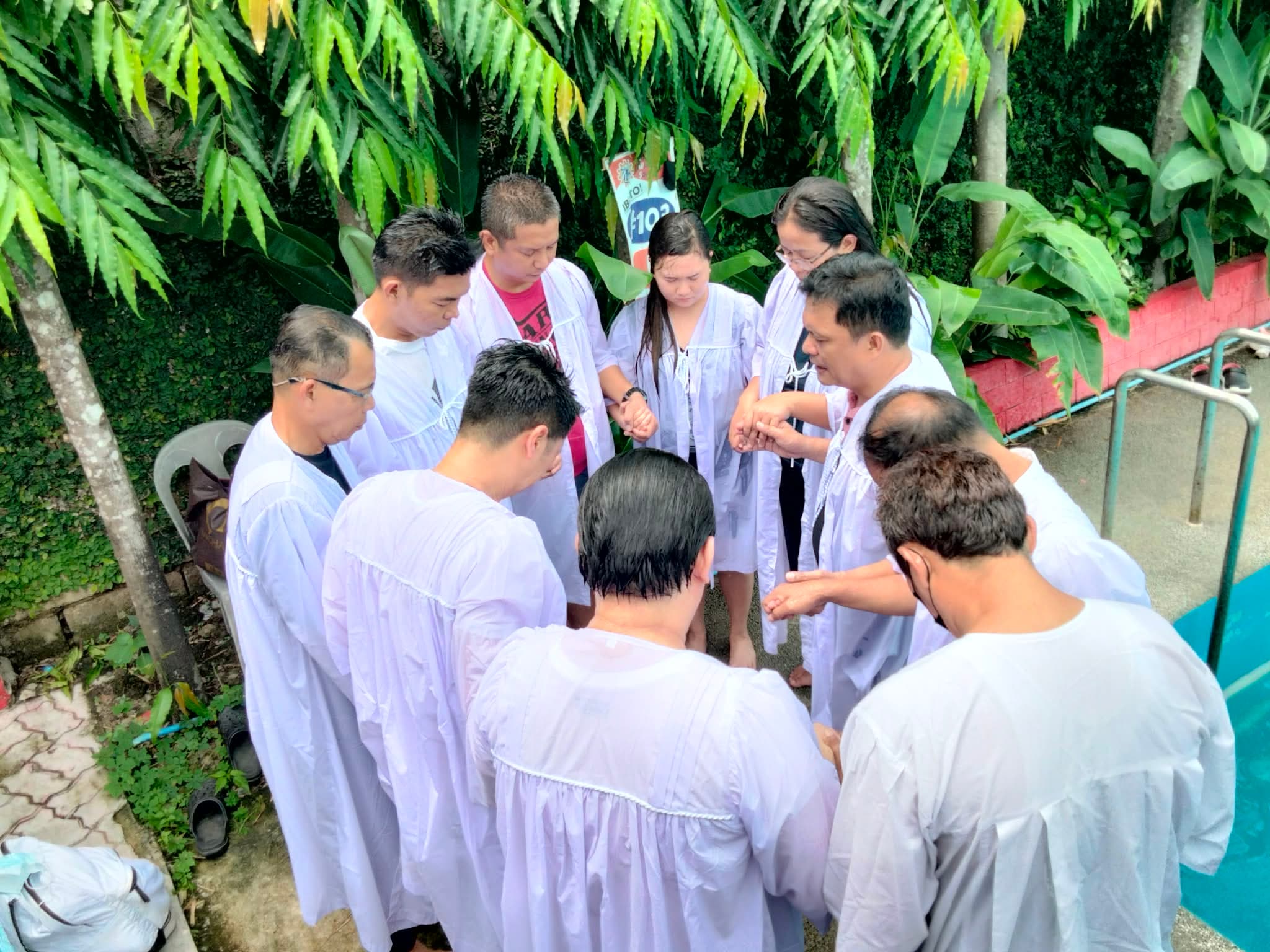 Baptism candidates and ministers in white robes praying together in a circle by the pool